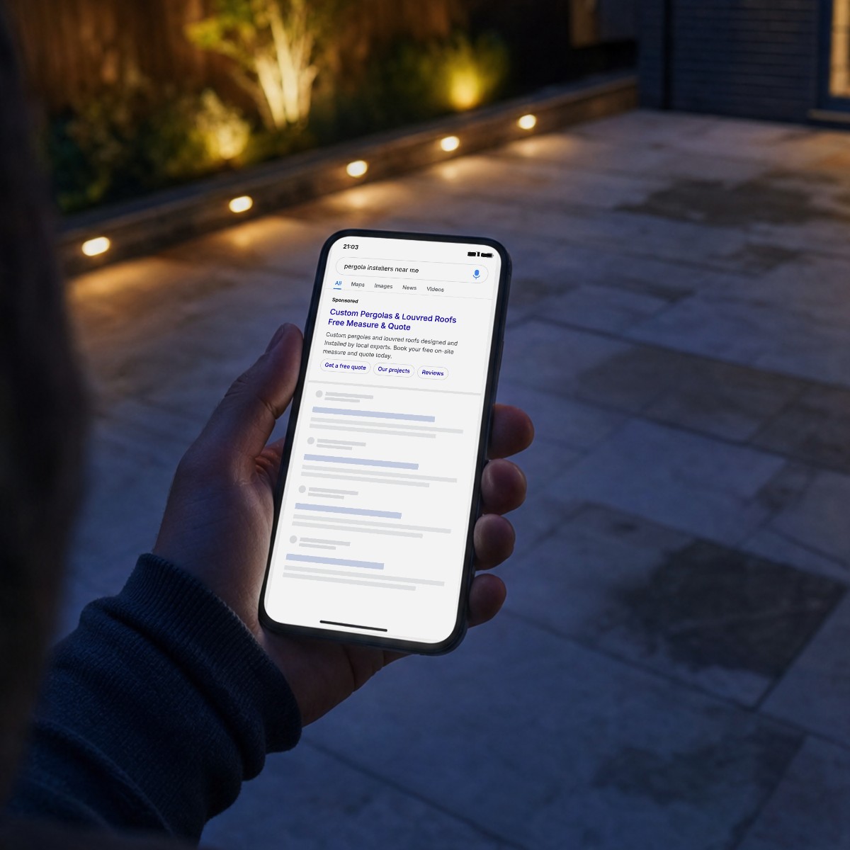 Homeowner on their patio at dusk searching for pergola installers near me, with a sponsored pergola ad at the top of the search results